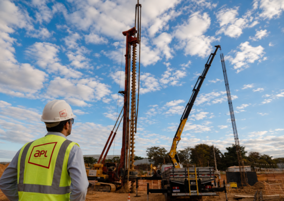 Engenheiro com uniforme da APL Engenharia observa execução de estaca hélice contínua monitorada em canteiro de obras, com perfuratriz, trado helicoidal e içamento de armadura.