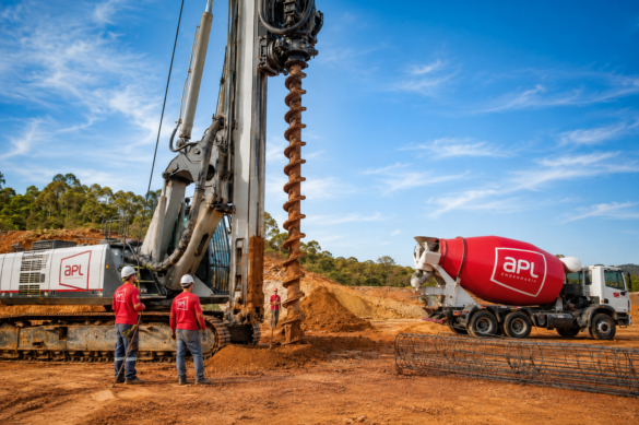 Equipamento de hélice contínua monitorada executando estaca de fundação em obra, com equipe da APL Engenharia, armaduras e caminhão betoneira com balão vermelho e logo oficial.