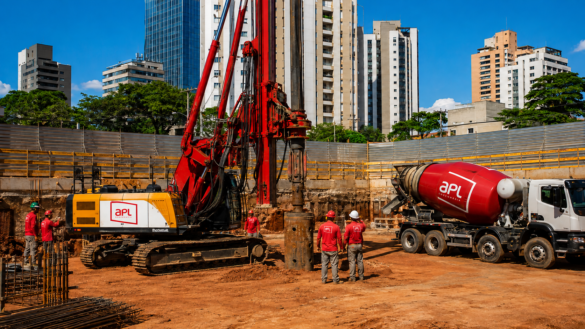 Perfuratriz e caminhão betoneira da APL Engenharia em obra urbana de fundações profundas, com equipe executando estacas e concretagem.