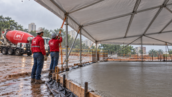 Trabalhadores com uniformes vermelhos da APL ajustam uma cobertura sobre laje de concreto, com caminhão-betoneira de balão vermelho ao fundo durante a chuva.